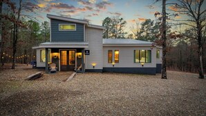 Une cabane moderne à deux étages, dotée d’un porche couvert et d’une allée en gravier, et entourée d’arbres.