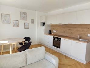Eine kompakte Küche mit weißen Schränken, einer Holzarbeitsplatte und einem eingebauten Ofen. Es gibt einen Essbereich mit Tisch und Stühlen, sowie ein Sofa im Wohnbereich.