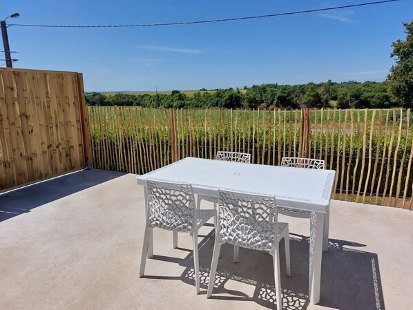 En udendørs terrasse med et hvidt bord og stole, omgivet af et træhegn og et marklandskab.