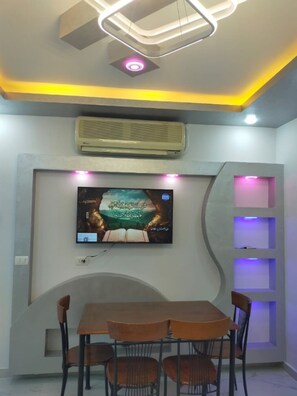 Un espace repas comprenant une table et des chaises, un climatiseur fixé au mur, un téléviseur mural et un design de plafond moderne avec des spots encastrés.