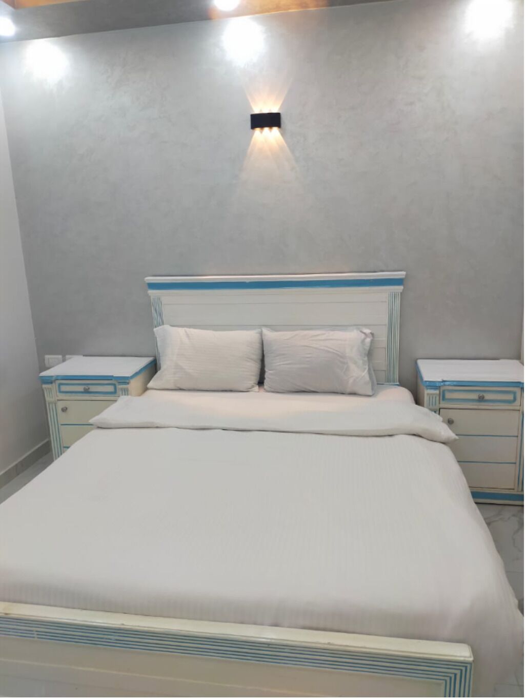 Un dormitorio con cama, dos mesitas de noche y una lámpara fijada a la pared.