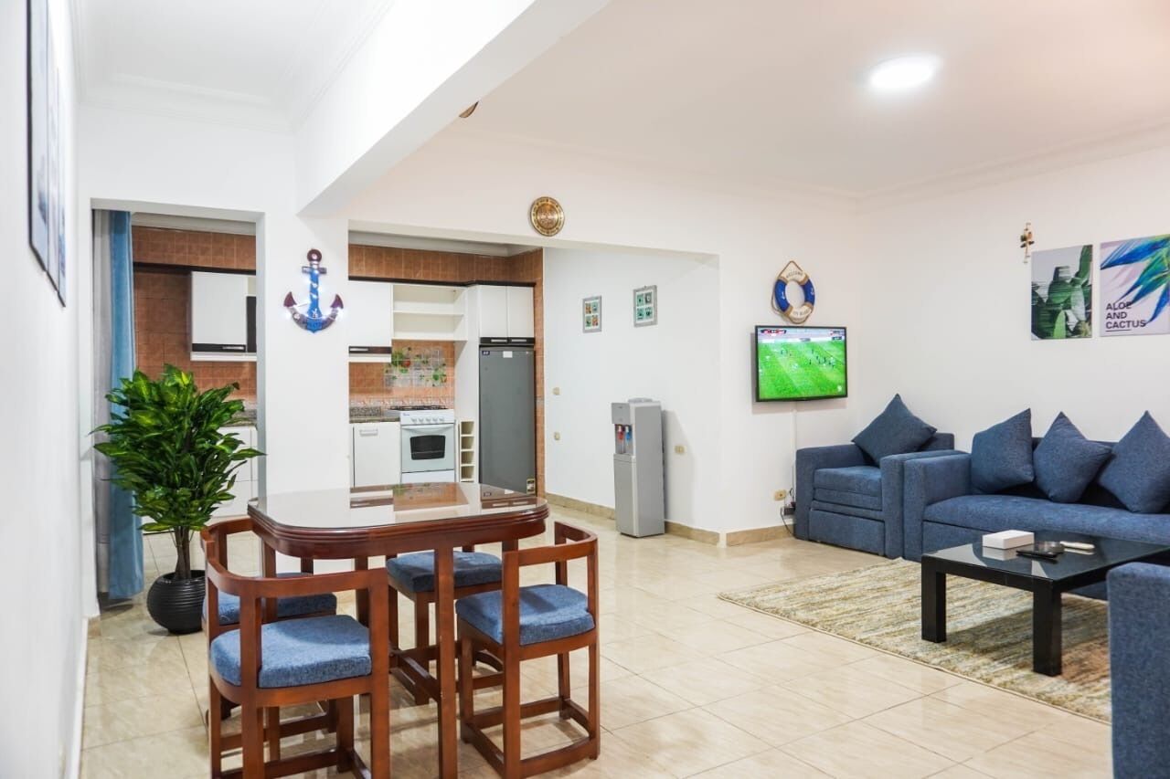 A modern living room with a blue sofa, a dining table with chairs, a TV mounted on the wall, and a kitchen area with cabinets and a refrigerator.