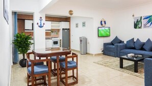 Een moderne woonkamer met een blauwe bank, een eettafel met stoelen, een aan de muur bevestigde televisie, en een keuken met kasten en een koelkast.