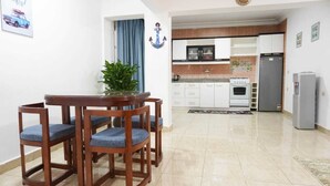 Een eetgedeelte met een houten tafel en stoelen, een kamerplant, en een keuken op de achtergrond met witte kasten en apparatuur.