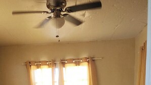 A bedroom with a large bed, a ceiling fan, a bedside table with a lamp, and curtains on the windows.