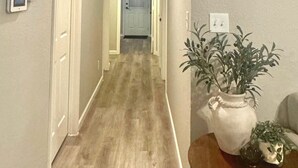 Un pasillo con piso de madera, una puerta blanca al final y una planta en maceta sobre una pequeña mesa.