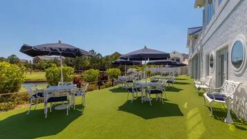 An outdoor patio with tables and chairs, surrounded by a pool and greenery.