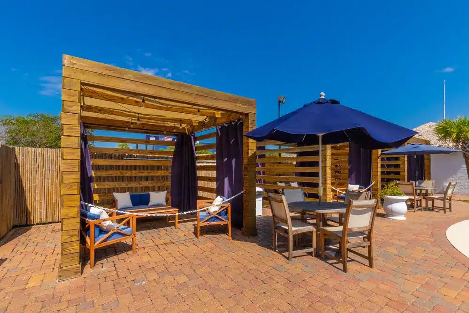 A wooden pergola with seating and umbrellas, brick flooring, and outdoor furniture.