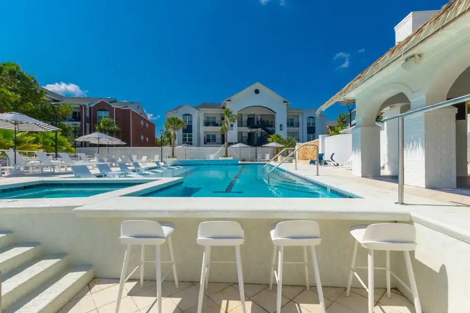 A swimming pool with two separate sections, a bar area with white stools, and a view of a large building with a white facade and arched windows.