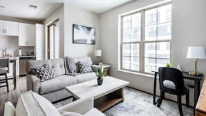 A modern living room with a grey sofa, a coffee table, and a black chair. There is a kitchen area with white cabinets and a window with a city view. - Landing Apartments near Rivergate Park Area (Cleveland)