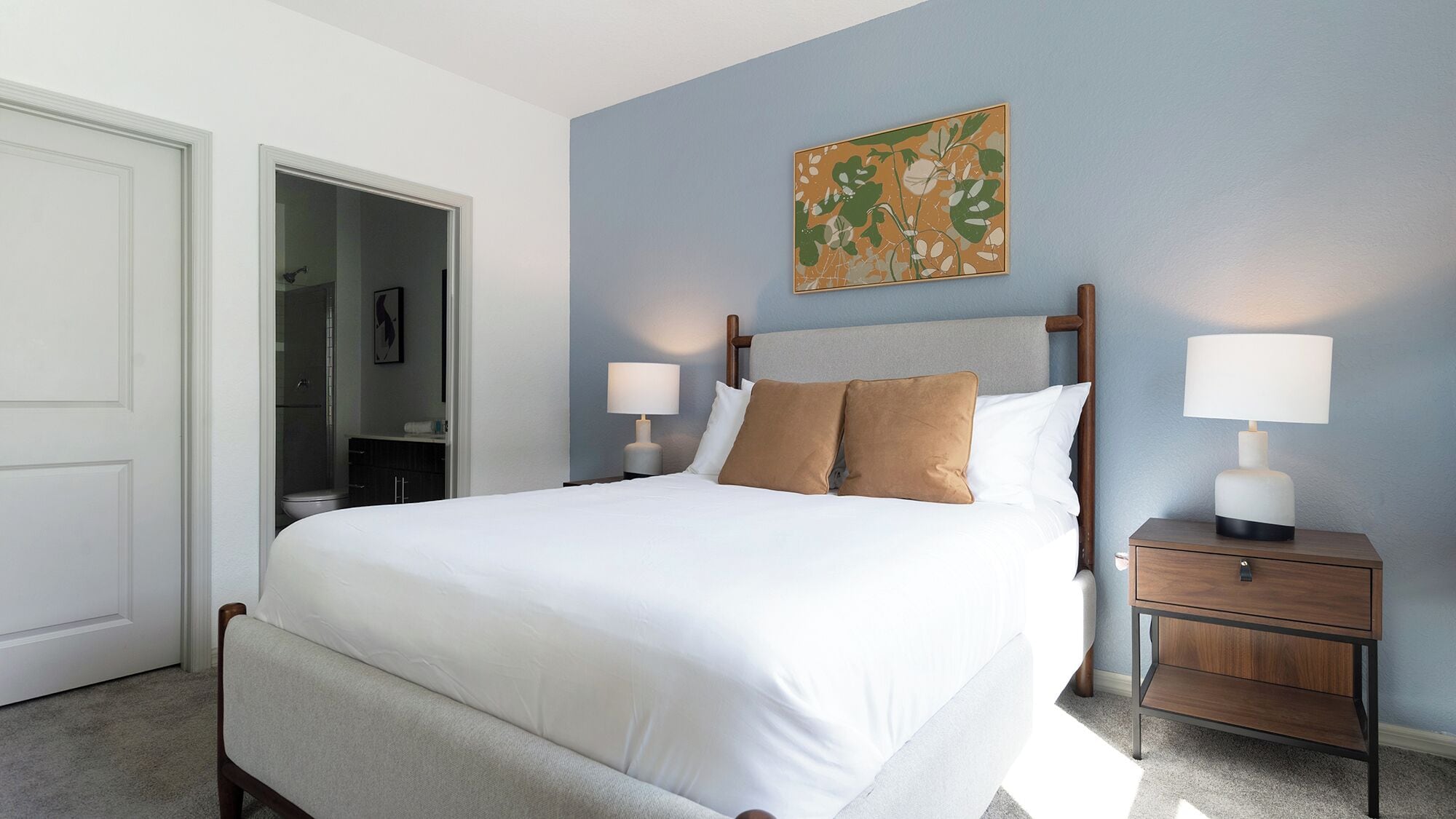 A neatly made bed with white linens and two brown pillows, a bedside table with a lamp, and a painting on the wall.