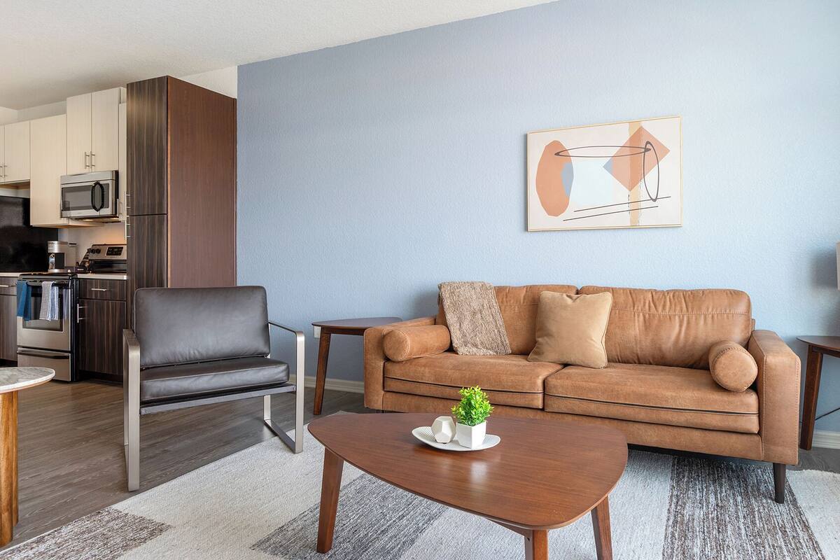A modern living room with a brown leather sofa, a black leather chair, a wooden coffee table, and a kitchen in the background.