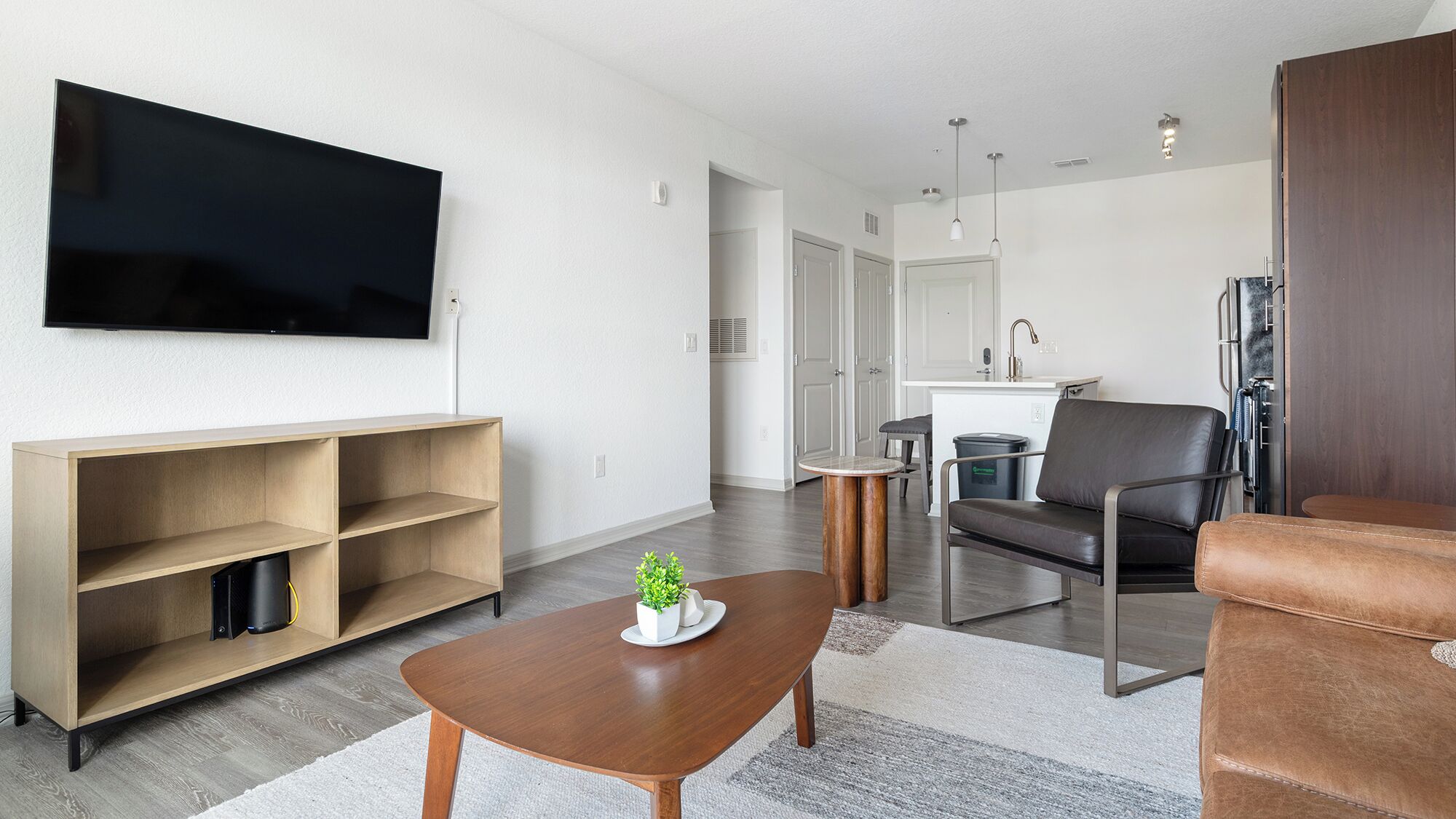 A modern living room with a flat-screen TV, wooden shelving, a coffee table, a leather sofa, and a kitchen area in the background.