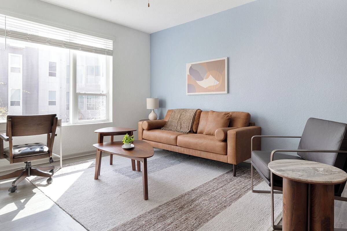 A modern living room with a brown leather sofa, a wooden coffee table, a side table with a lamp, a grey armchair, and a large window with blinds.