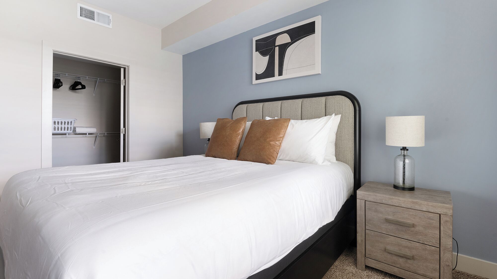 A neatly made bed with white linens, a headboard with a grey fabric panel, and two brown pillows. There is a bedside table with a lamp and a small drawer. A framed abstract artwork hangs on the wall above the bed.