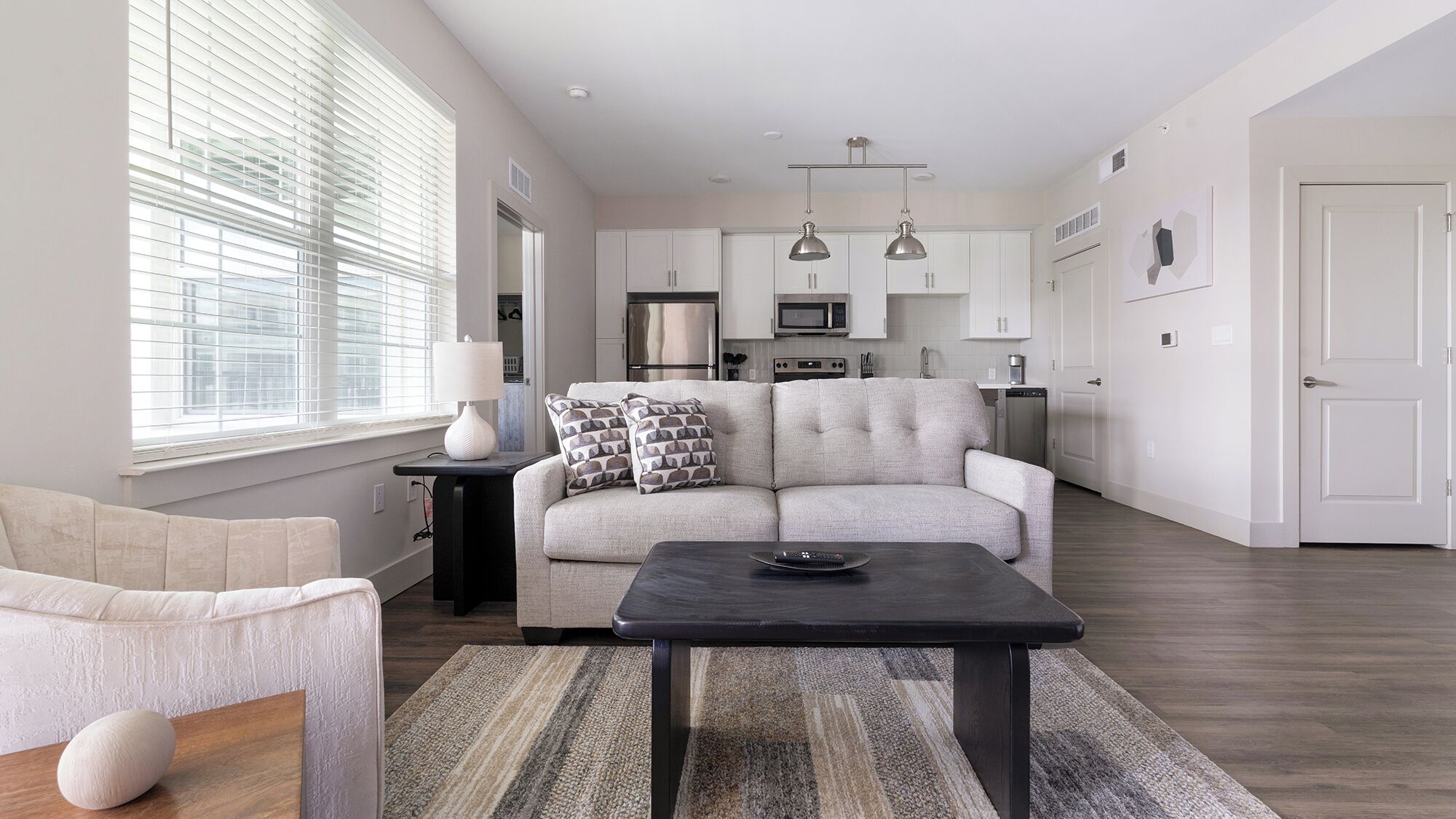 A modern living room with a sofa, a coffee table, and a lamp. There is a kitchen area with cabinets and appliances in the background.