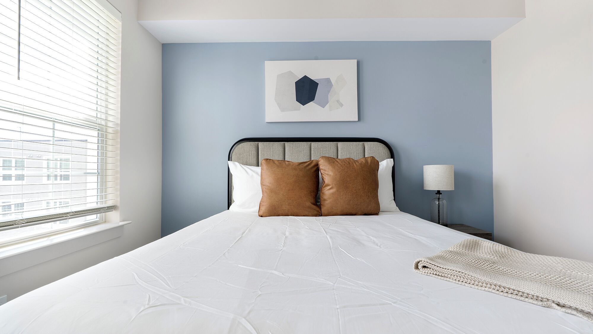Un lit bien fait, avec du linge de lit blanc et deux oreillers marron, une lampe de chevet et une œuvre d’art abstraite encadrée accrochée au mur.
