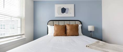 Un lit bien fait, avec du linge de lit blanc et deux oreillers marron, une lampe de chevet et une œuvre d’art abstraite encadrée accrochée au mur.