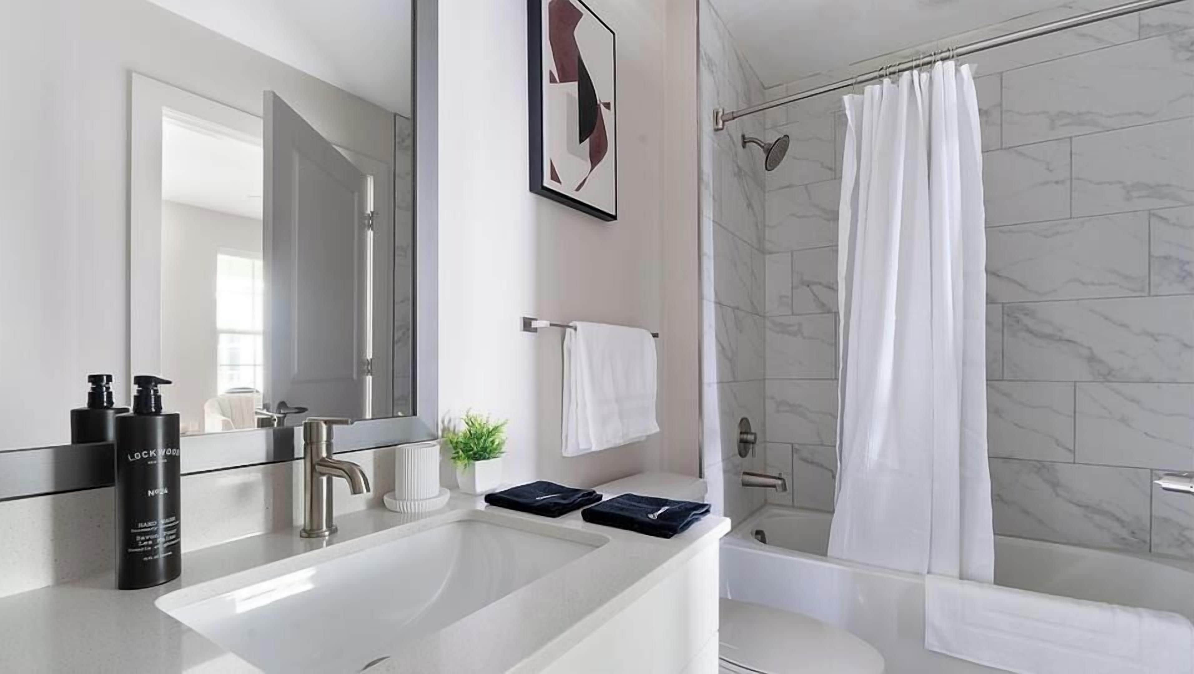 Een moderne badkamer met een grote spiegel, een witte wastafel, twee zwarte flessen, een handdoekhouder en een douche met een witte douchegordijn.