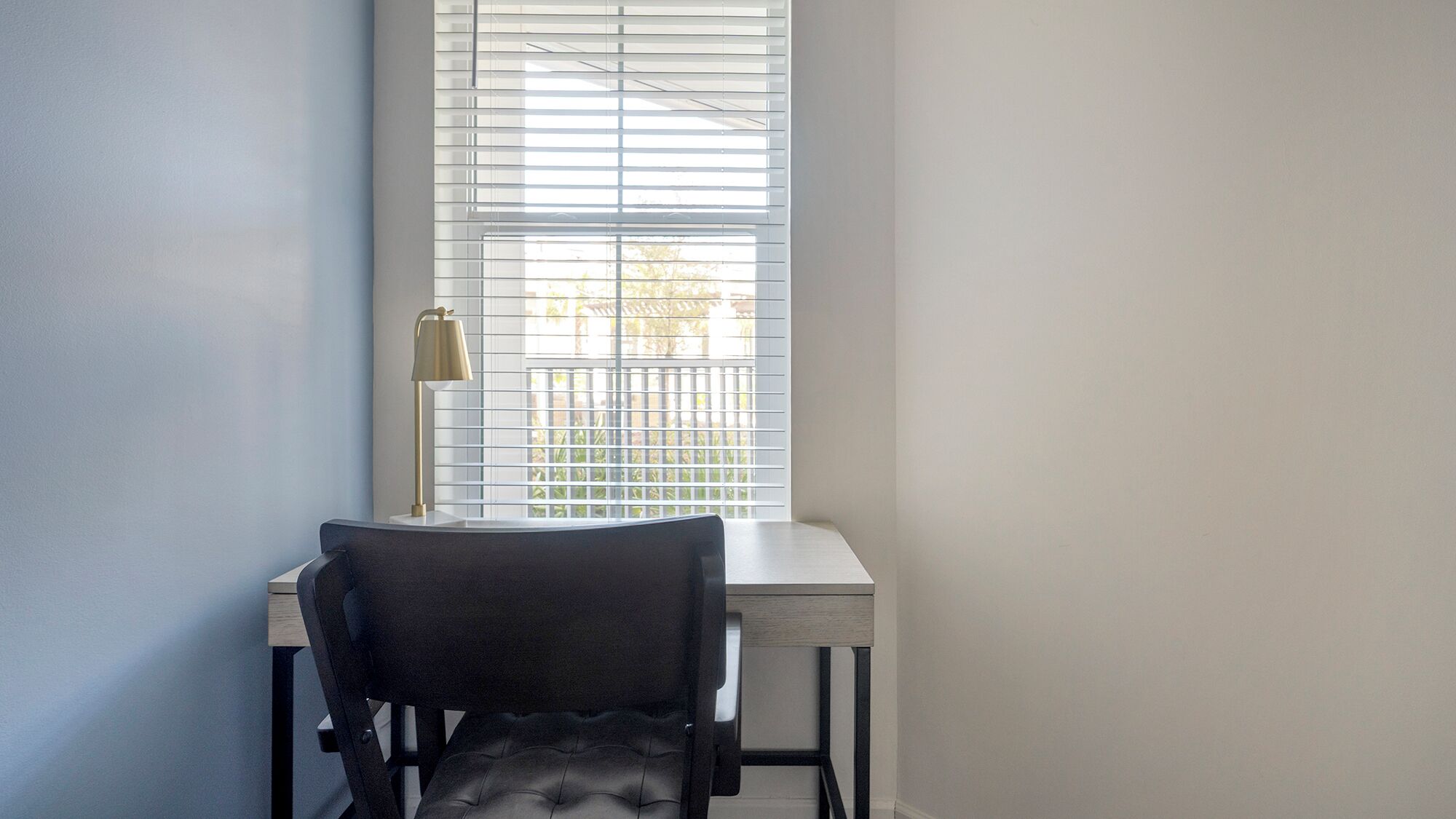 A corner room with a desk and chair, a window with blinds, and a lamp.