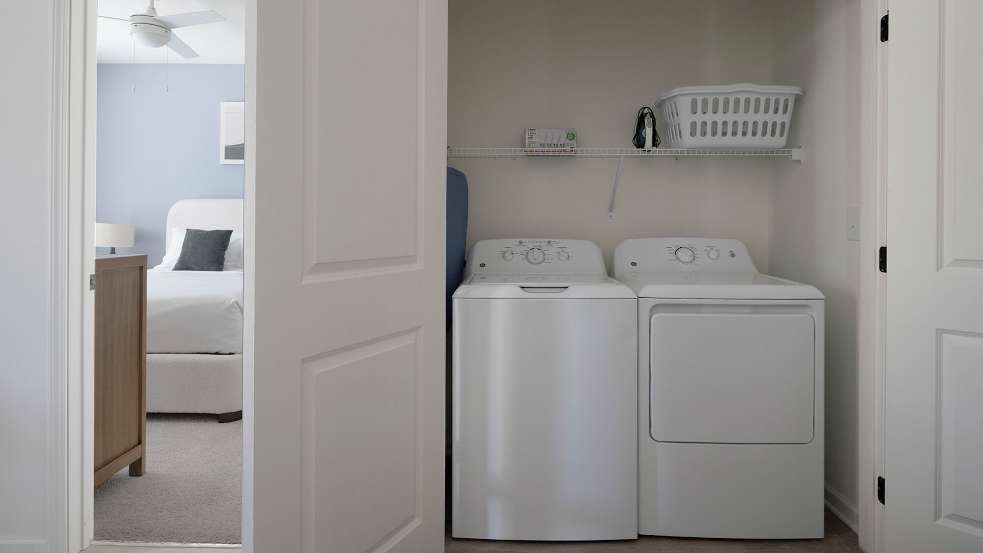 Uma lavanderia com duas máquinas de lavar e secar de carregamento frontal brancas, uma prateleira com cestos de roupa, e uma cama no quarto adjacente.