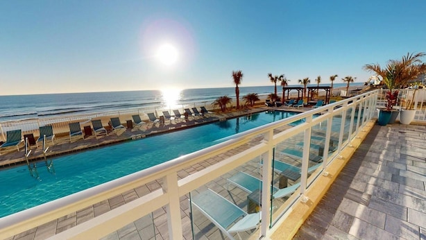 Blick auf einen Pool mit blauem Himmel, sonnigem Meer und einer Holzterrasse mit Liegestühlen.