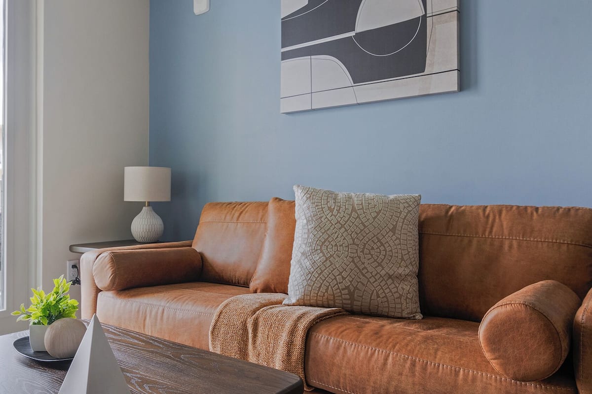 Una sala de estar moderna con un sofá de cuero marrón, un cojín decorativo estampado, una mesita auxiliar con una lámpara y vistas de la ciudad a través de un ventanal grande.