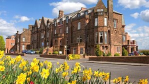 A historic brick building with multiple windows and a prominent corner turret, situated on a street with daffodils in the foreground. - Cairndale Hotel & Spa (Dumfries)