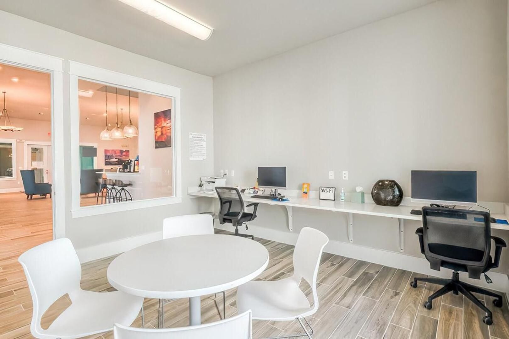 Een moderne kantoorruimte met een ronde tafel, witte stoelen, twee bureaus met computers en een groot raam.