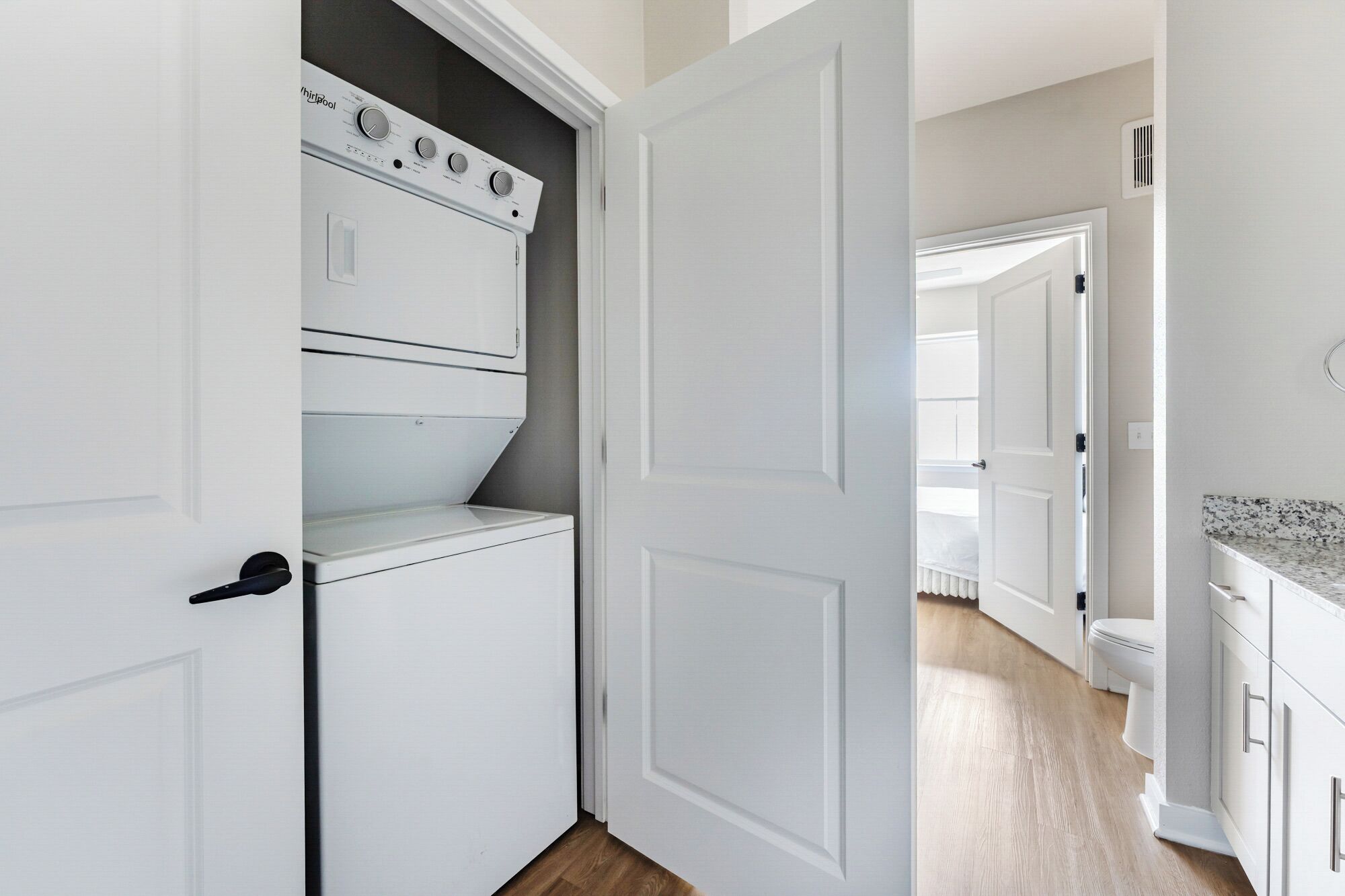 Une buanderie équipée d’un lave-linge et d’un sèche-linge superposés, une porte donnant sur une chambre, et un coin cuisine avec un plan de travail.