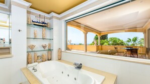 Une salle de bain moderne avec une grande baignoire, des étagères en verre ornées d’objets décoratifs et une vue sur un patio extérieur avec une table et des chaises.