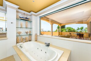 Ein modernes Badezimmer mit einer großen Badewanne, Glastablaren mit Dekoration und Blick auf eine Terrasse mit Tisch und Stühlen.