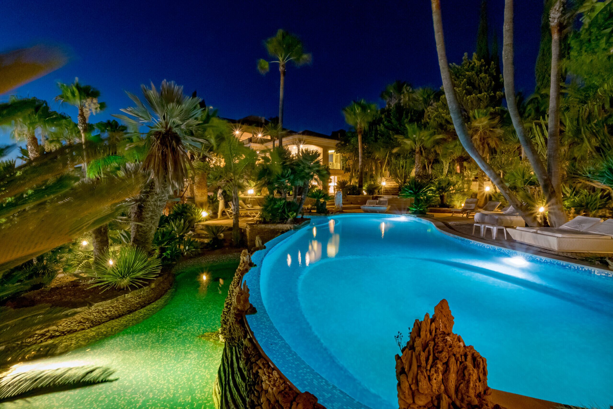 A nighttime view of a resort pool surrounded by palm trees and a well-lit building.