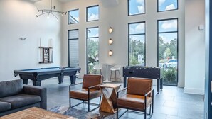 A spacious living area with a pool table, two leather chairs, a coffee table, and a foosball table. Large windows provide natural light and views of the outdoors. - Landing Apartments Near North Okaloosa Medical Center (Crestview)