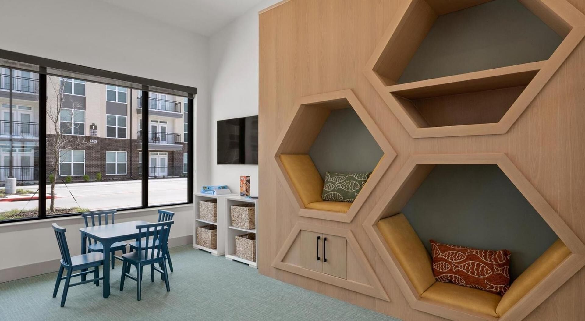 A modern room with a large window, a dining area with blue chairs, and a unique hexagonal shelving unit with cushions.
