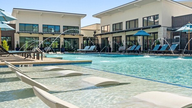 A swimming pool with lounge chairs and umbrellas, surrounded by modern buildings with large windows. - Landing Furnished Apartments - Garland (Sachse)