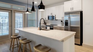 A modern kitchen with white cabinets, a stainless steel refrigerator, a microwave, and a large island with stools. - Landing Furnished Apartments - Garland (Sachse)