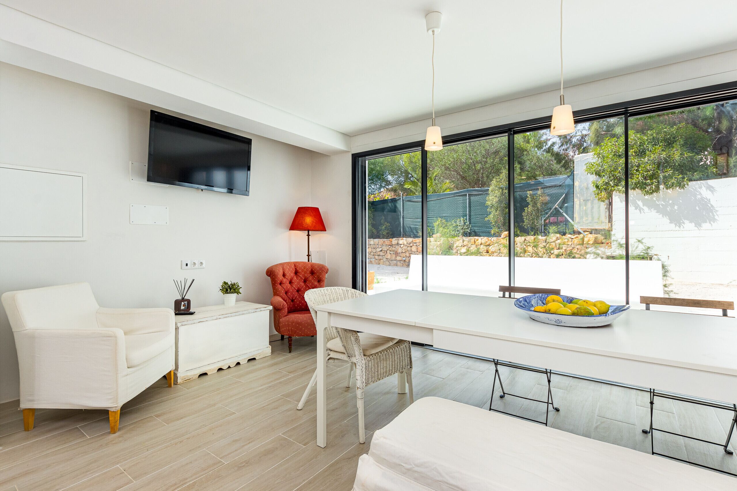 Een moderne woonkamer met een witte bank, een rode fauteuil, een eettafel met een fruitschaal, en een aan de muur bevestigde televisie.