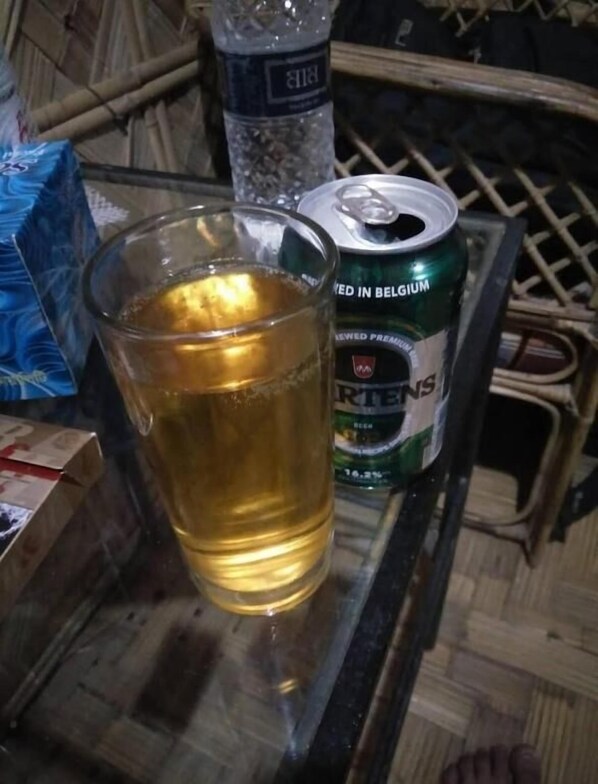 A glass of beer next to a can of Pilsner beer on a table with a woven basket and a plastic bag in the background. - Nishorgo Nirob Eco Resort (Kamalganj)