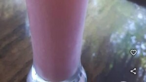 Un grand verre contenant une boisson rose et une paille verte, posé sur une surface réfléchissante avec un arrière-plan extérieur flou.