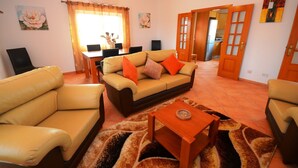 Ein Wohnzimmer mit einer Couch, zwei Sesseln, einem Couchtisch und einem Essbereich mit Tisch und Stühlen. Durch eine offene Tür ist eine Küche zu sehen.