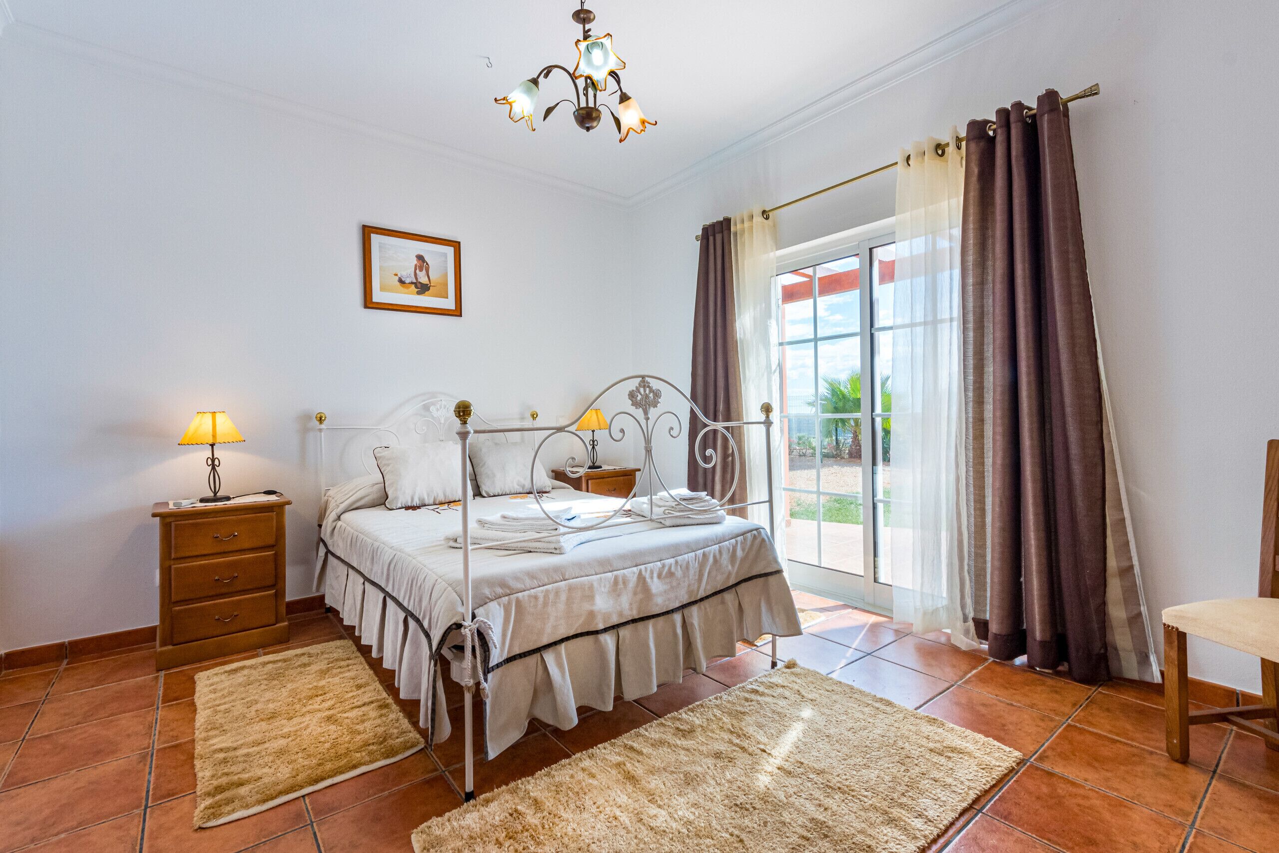 A bedroom with a four-poster bed, a nightstand, a lamp, a chair, and a view of the outdoors through a large window with curtains.