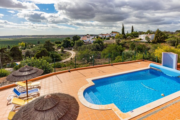 A swimming pool with a curved edge, surrounded by lounge chairs and umbrellas, with a view of houses and greenery in the background. - Villa da Horta, a beautiful extensive villa in a peaceful location (Alcantarilha)