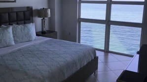 Un dormitori amb un llit gran, una tauleta de nit, una làmpada i vista a l'oceà a través d'una finestra ampla.