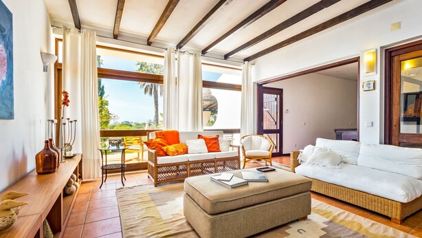 A spacious living area with a sofa, ottoman, and a dining table with chairs. There is a large window providing a view of the outdoors.