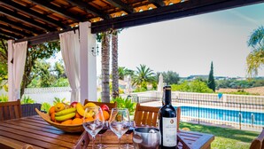 A wooden table with a fruit bowl, wine glasses, and a bottle of wine, overlooking a pool and greenery.
