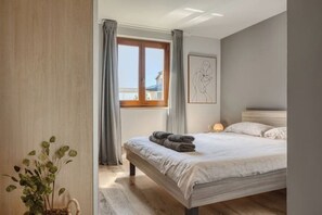Ein Schlafzimmer mit einem großen Bett, einem Fenster mit Vorhängen, einem Nachttisch mit Lampe und einem gerahmten Kunstwerk an der Wand.