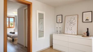 Un corridoio con un letto sullo sfondo, una cassettiera bianca con opere d'arte incorniciate e uno specchio appeso al muro.