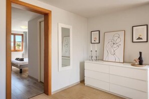 Ein Flur mit einem Bett im Hintergrund, einer weißen Kommode mit gerahmten Bildern und einem Spiegel an der Wand.