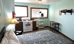 Une pièce compacte comprenant un lit, une kitchenette avec une cuisinière et un four, une petite table et une fenêtre avec des stores.